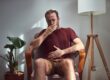 Man sits in a living room chair yawning with his hand over his mouth beneath a maroon shirt.