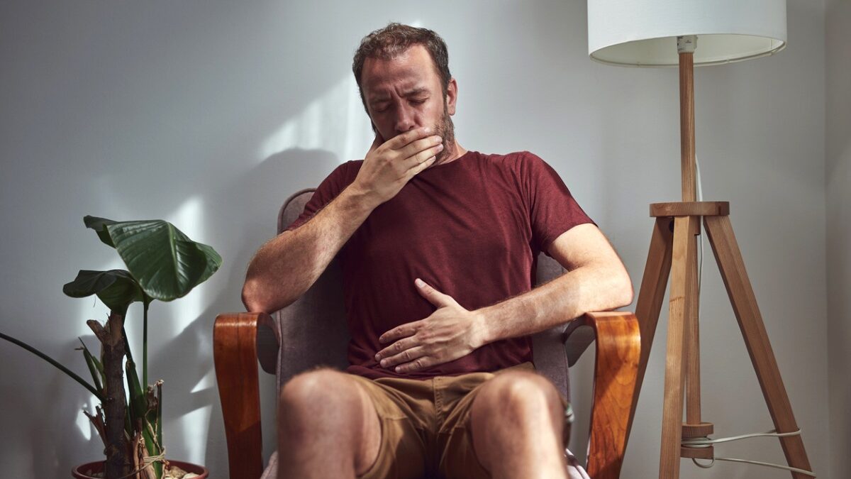Abdominal Pain and Vomiting: Stomach Bug or Appendicitis? Man sits in a living room chair yawning with his hand over his mouth beneath a maroon shirt.