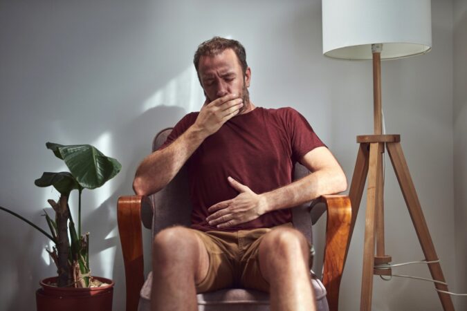 Man sits in a living room chair yawning with his hand over his mouth beneath a maroon shirt.
