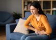 Woman sits on a blue couch, grimacing in pain while clutching her abdomen at home.