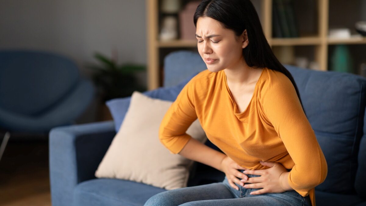 Woman sits on a blue couch, grimacing in pain while clutching her abdomen at home.