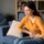 Woman sits on a blue couch, grimacing in pain while clutching her abdomen at home.