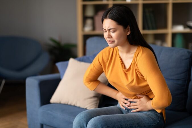 Woman sits on a blue couch, grimacing in pain while clutching her abdomen at home.