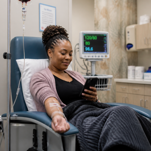 Woman seated in a hospital bed with an IV in her arm, using a smartphone while bundled under a blanket; patient monitor with vital signs visible in the background.